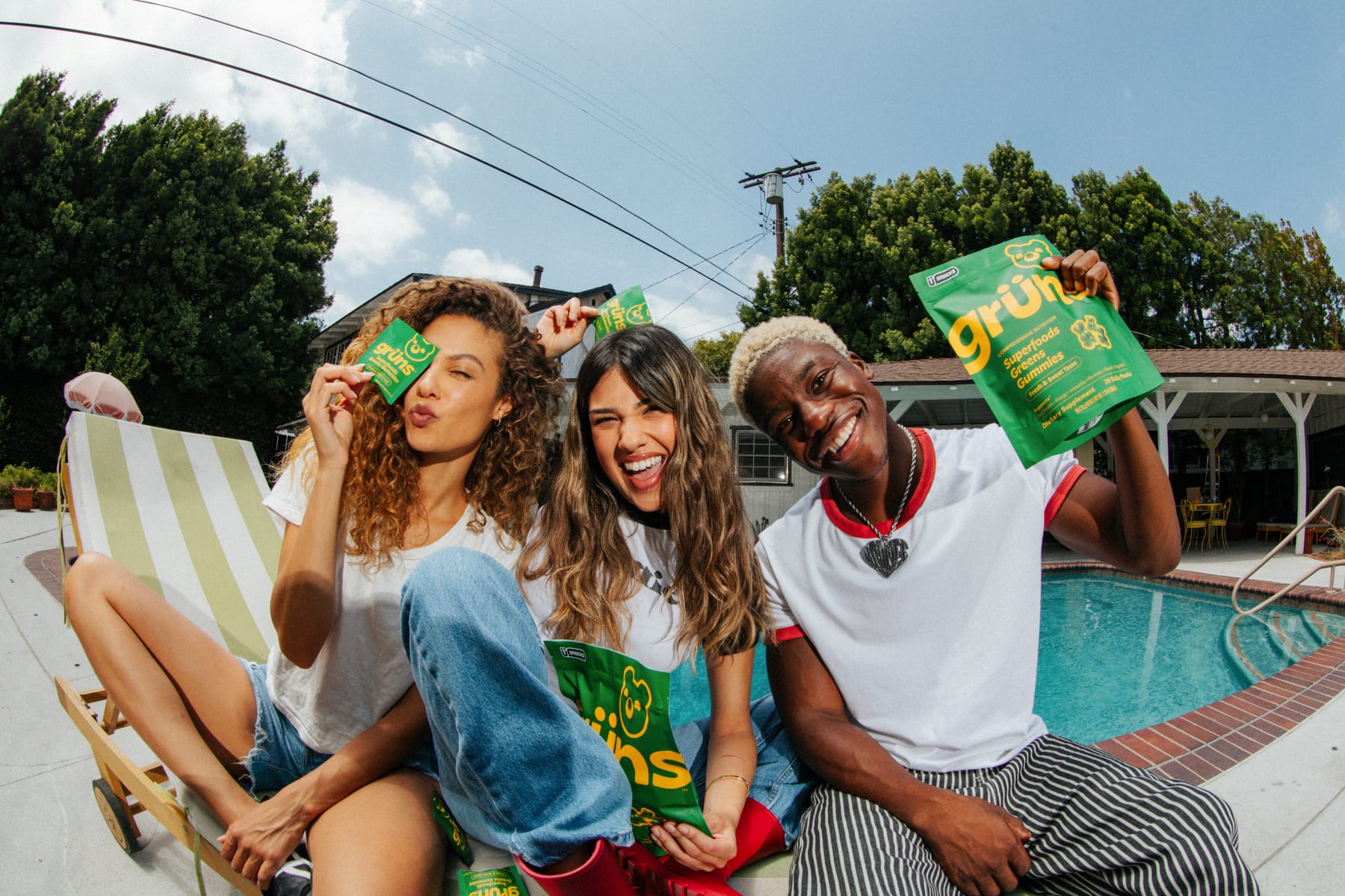 Three friends laughing by a pool, holding up Grüns Superfoods Greens Gummies packs