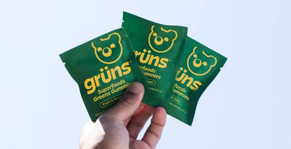 A hand holding up three Grüns Superfoods Greens Gummies snack packs against a pale sky